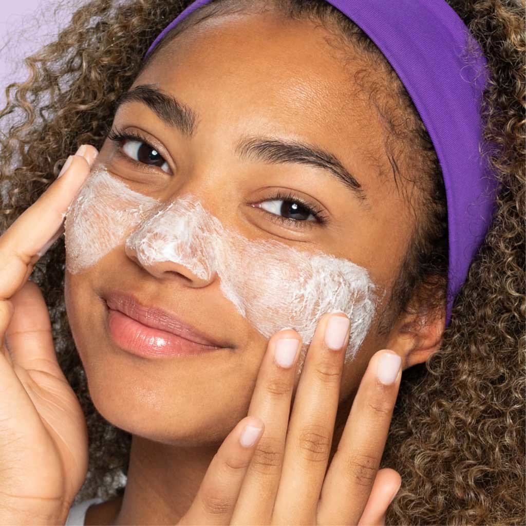 Young brown eye teen with purple headband and curls applying deep action cream cleanser for sensitive skin to cheeks