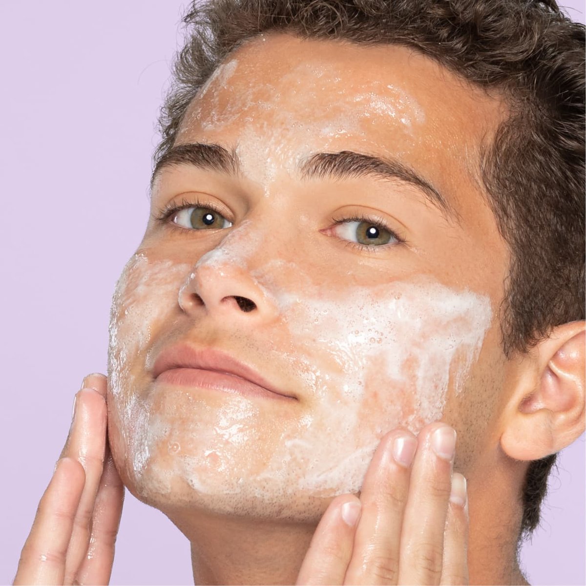 Young teen with hazel eyes and brown hair applies foaming facial cleanser for sensitive skin with hands on cheek in front of light purple background