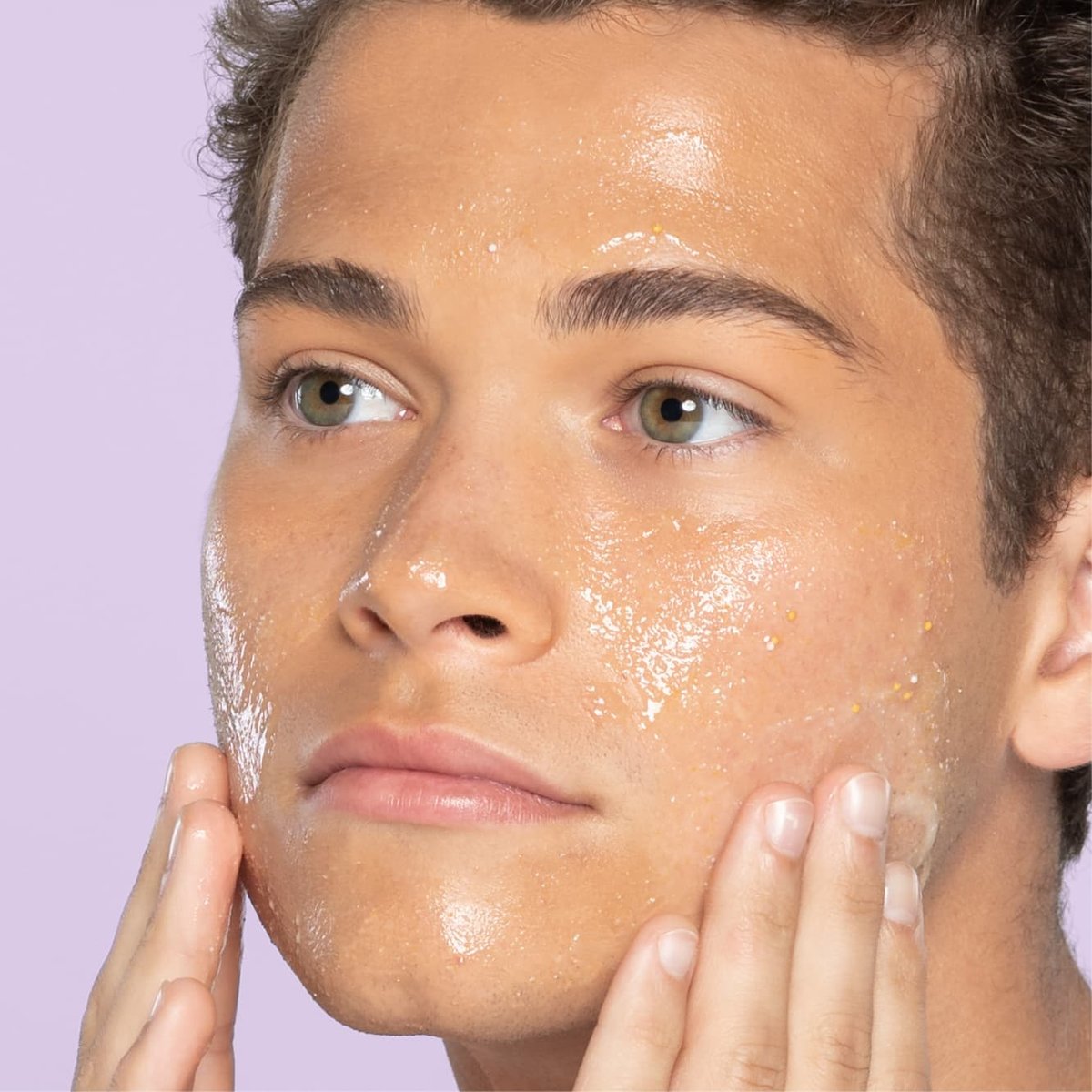 Young teen with hazel eyes and light brown hair applies lemon zesty scrub to cheeks in front of light purple background