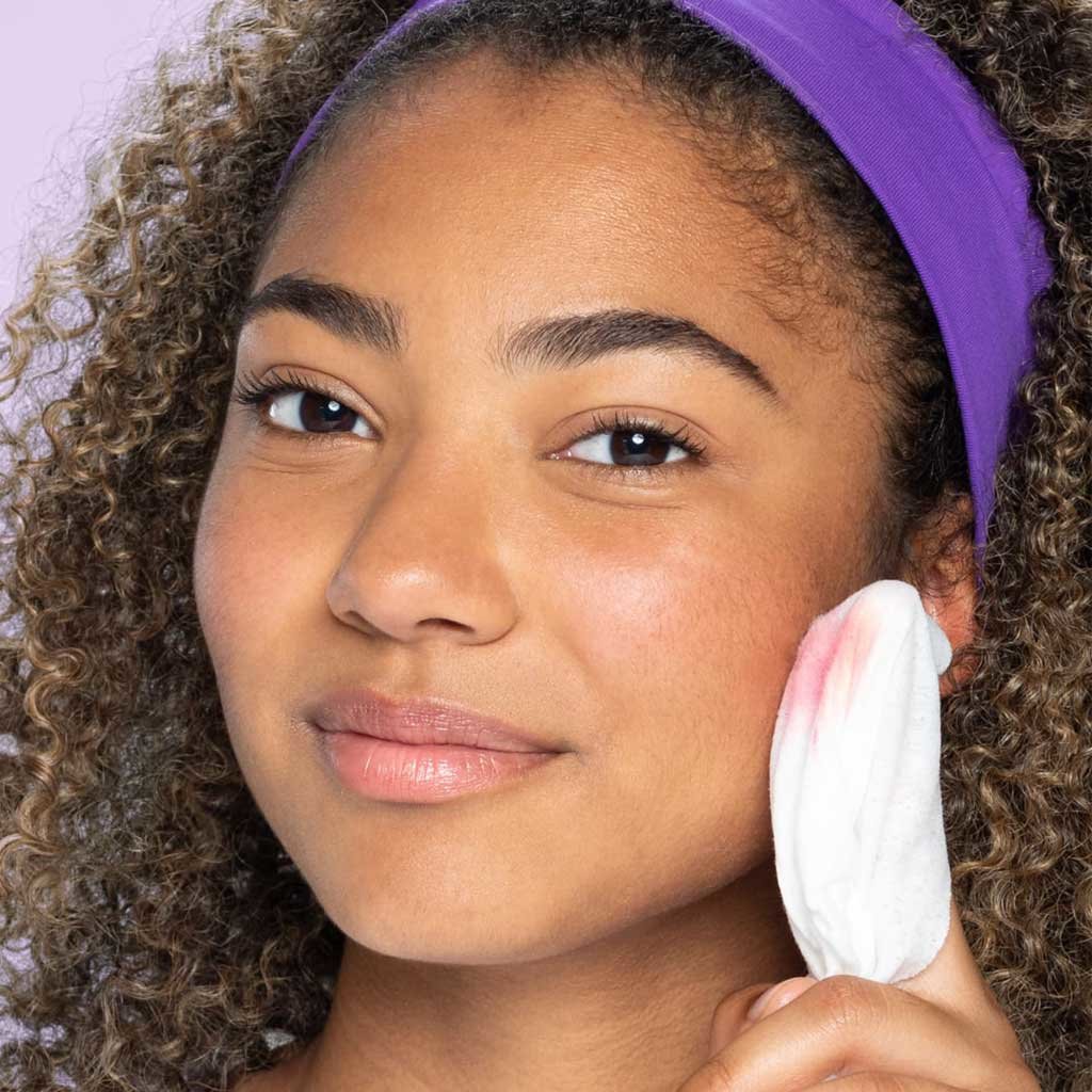 Young teen with brown eyes brown and curly hair with a purple headband wipes blush off cheek with Clean & Clear makeup dissolving cleansing wipe