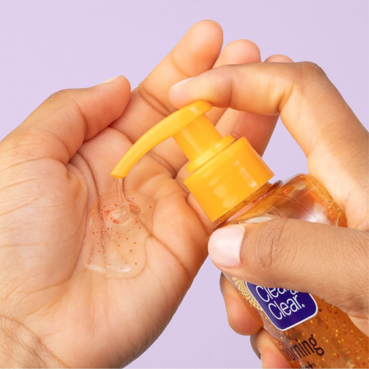 Orange Clean & Clear Morning Burst cleanser being pumped into palm of hand in front of purple background