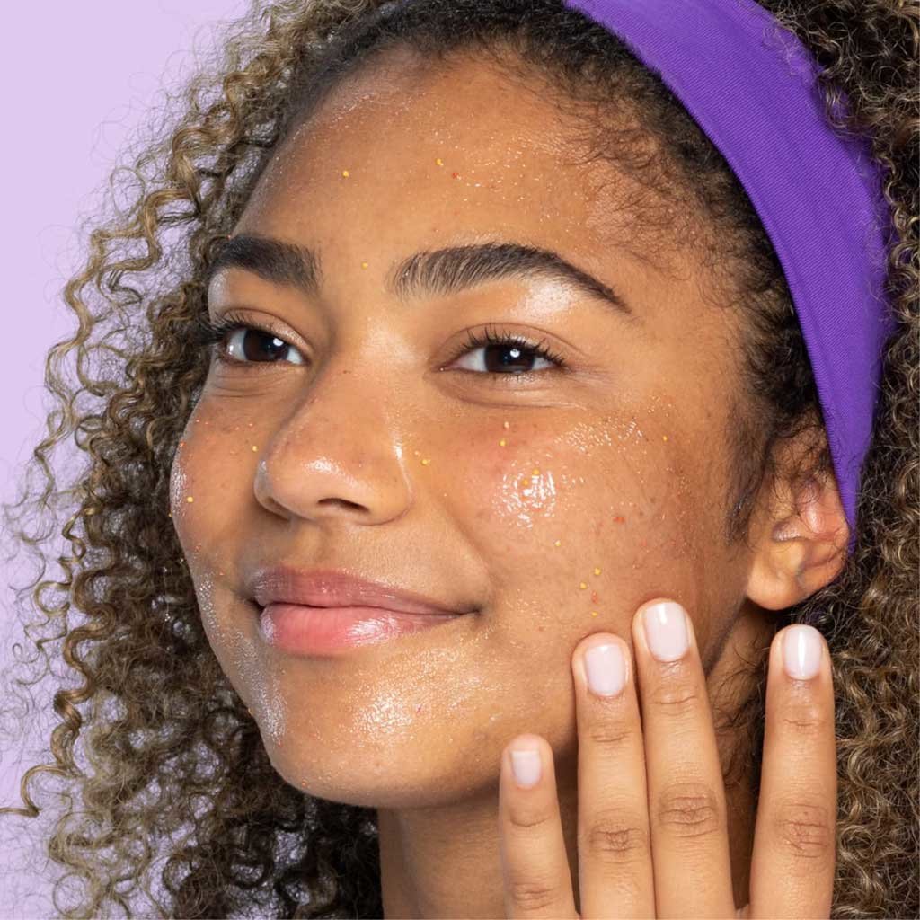 Young brown eye teen with purple headband and curls applying daily pore cleanser to cheeks