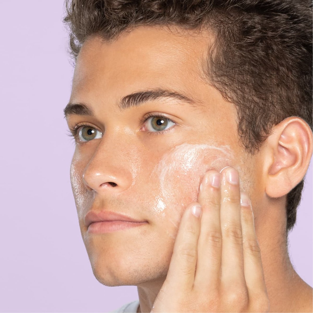 Young teen with hazel eyes and light brown hair applies foaming Night Relaxing face wash to cheek in front of light purple background