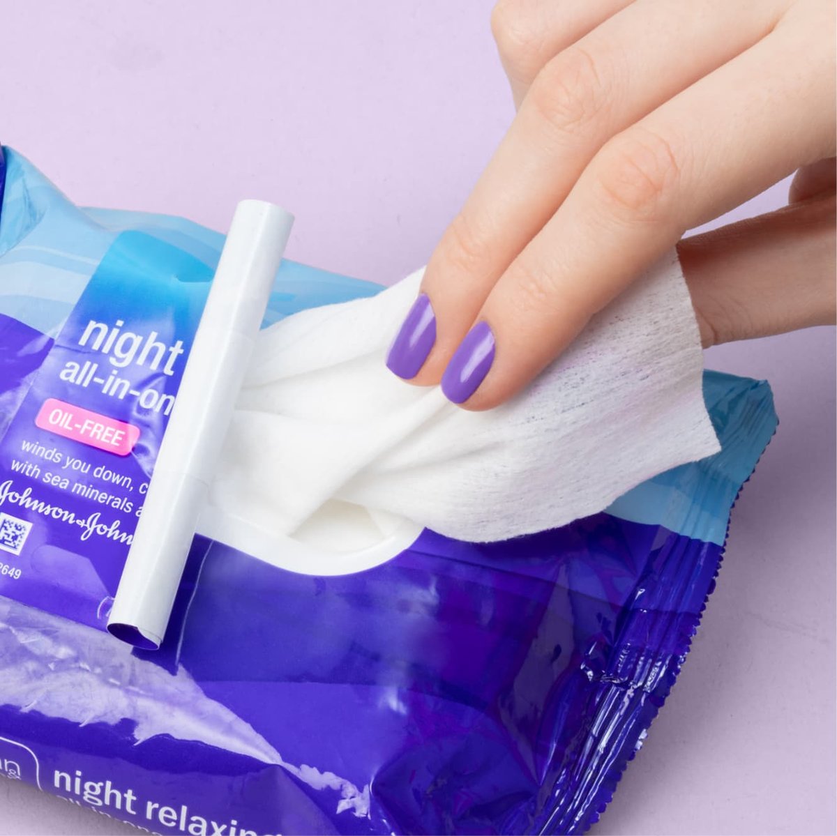 Hand with purple nails pulls Night Relaxing cleansing wipe out of dark blue reusable packaging in front of light purple background