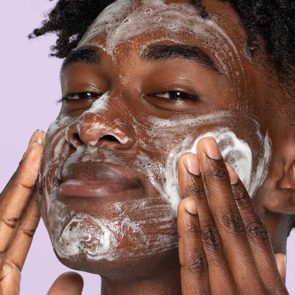 Young teen with brown eyes and dark brown twists washes his cheeks with foaming watermelon gel cleanser in front of light purple background