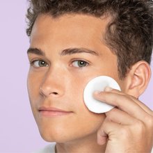 Young teen with hazel eyes and brown hair applies deep cleaning toner for sensitive with a pad on cheek in front of light purple background